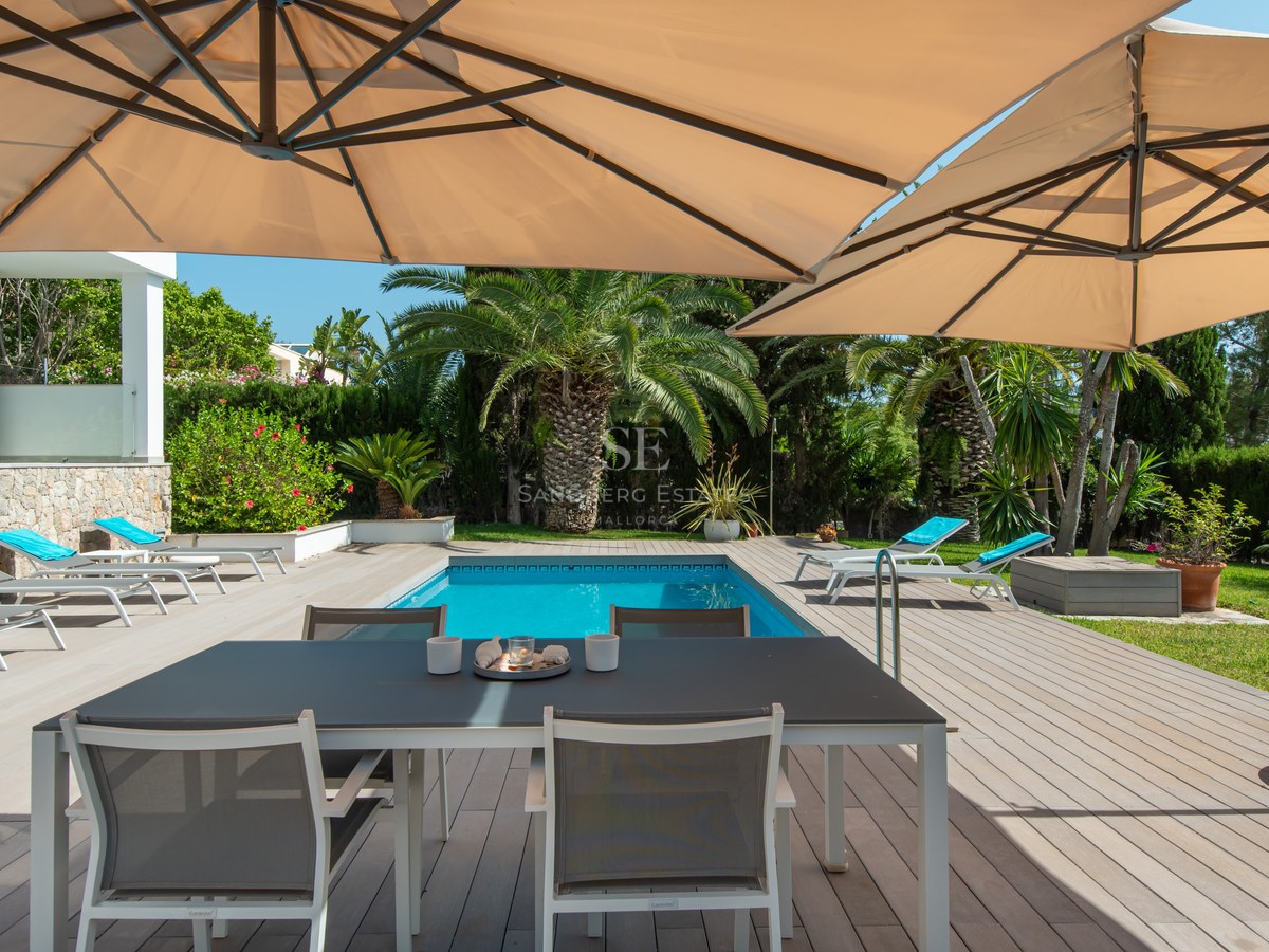 Wooden deck terrace featuring a dining table under large umbrellas next to a swimming pool and tropical palm trees.