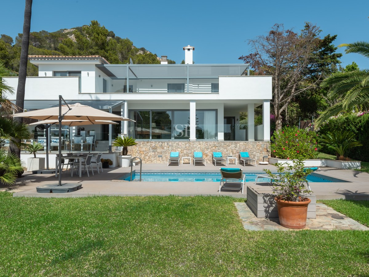 White contemporary villa featuring a swimming pool, sun loungers, and lush green lawn under a clear blue sky.