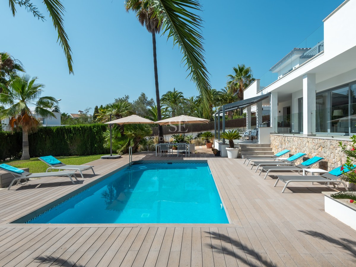 Rectangular swimming pool surrounded by wooden decking, white sun loungers, and palm trees under a clear blue sky.