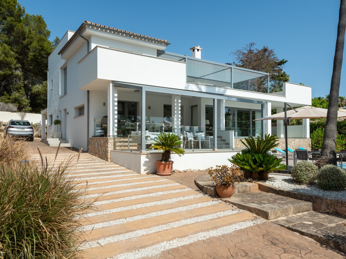 Two-story modern white villa with glass-fronted terraces, stone accents, and manicured landscaping under a clear blue sky.