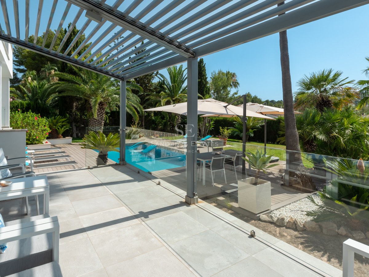 Modern covered terrace with lounge furniture overlooking a swimming pool and lush palm garden under a clear blue sky.