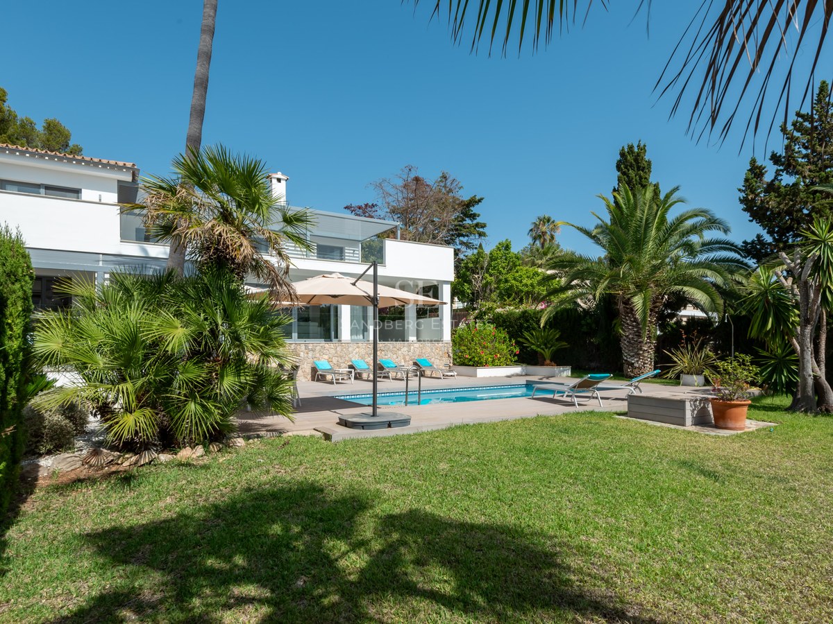 Modern white villa with a turquoise swimming pool, sun loungers, and lush palm trees under a clear blue sky.
