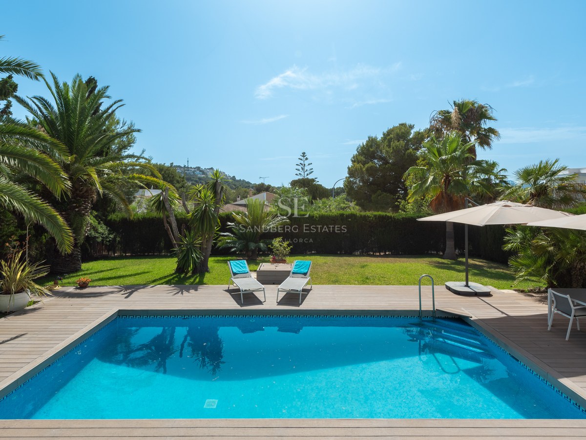 A rectangular turquoise pool surrounded by a wooden deck, sun loungers, and lush palm trees under a bright blue sky.
