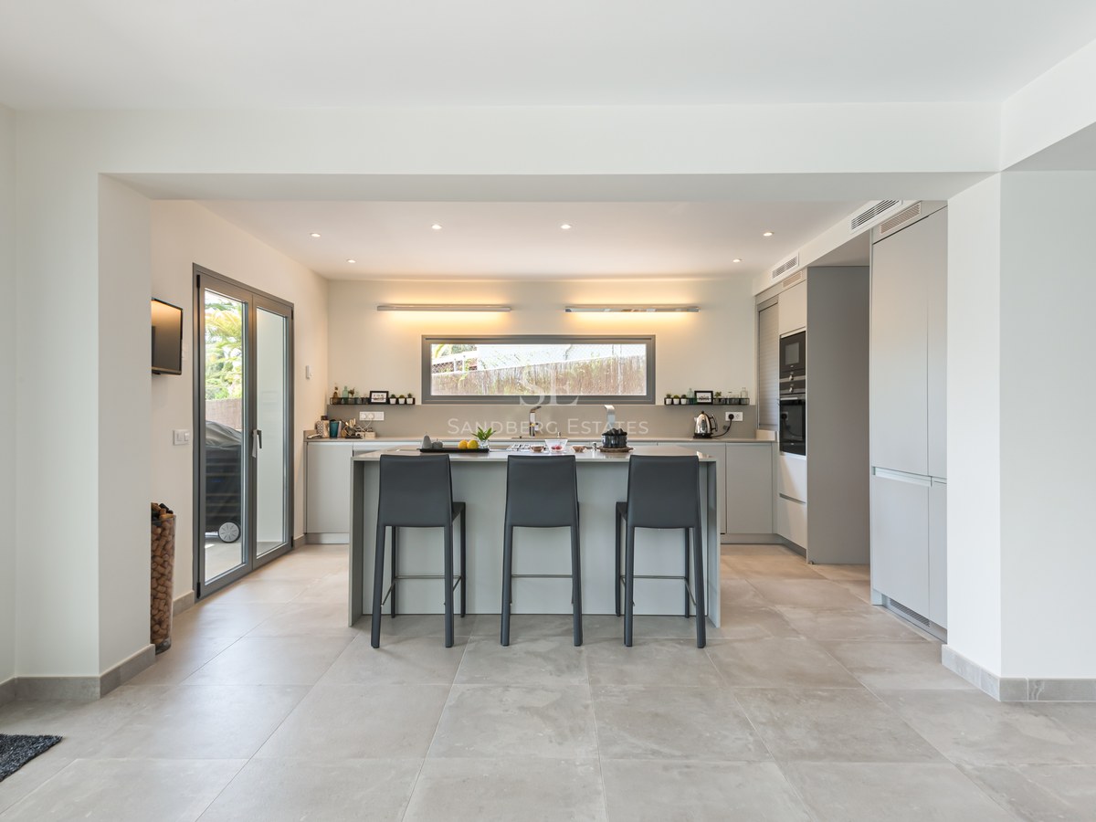A minimalist grey and white kitchen featuring a large central island with charcoal bar stools and porcelain tile floors.