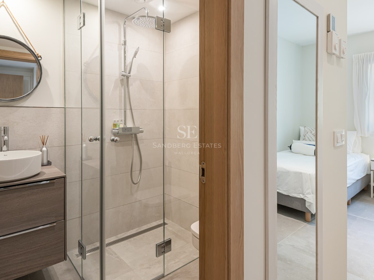 Modern bathroom featuring a glass walk-in shower, ceramic vessel sink, and round mirror, looking into a bedroom.