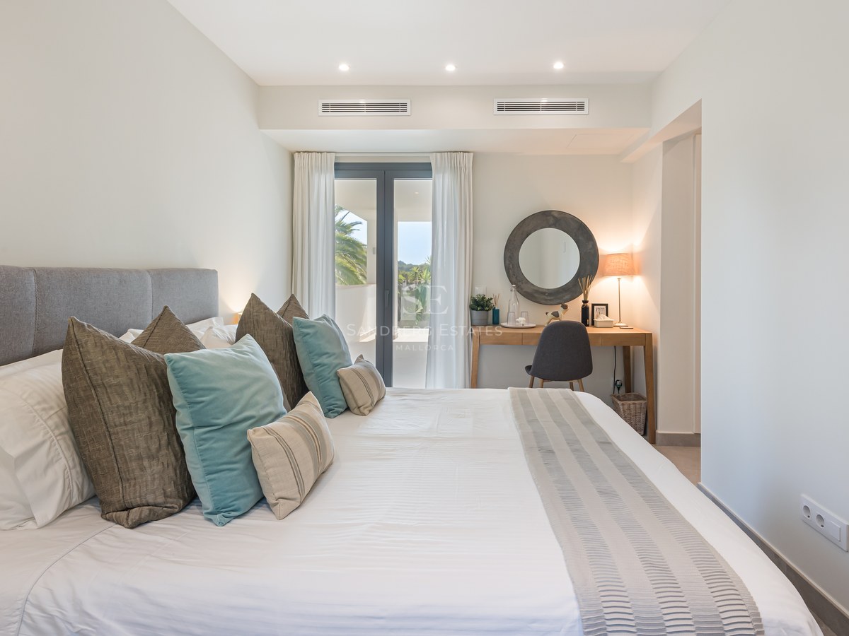 Bright bedroom featuring a large bed with teal accents, a wooden vanity desk, and floor-to-ceiling glass doors leading to a terrace.