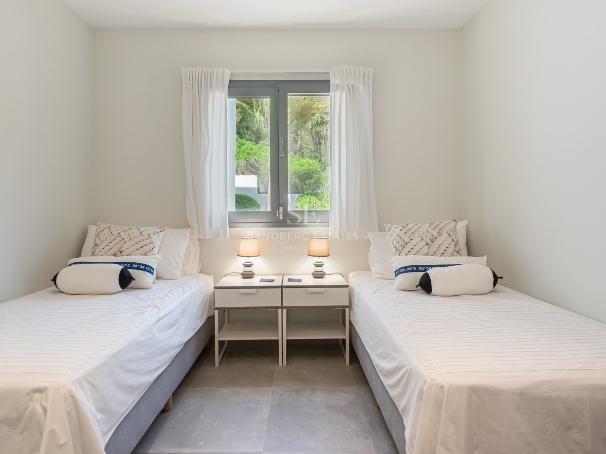 Bright guest bedroom featuring two single beds, white linens, and a window overlooking lush green gardens.