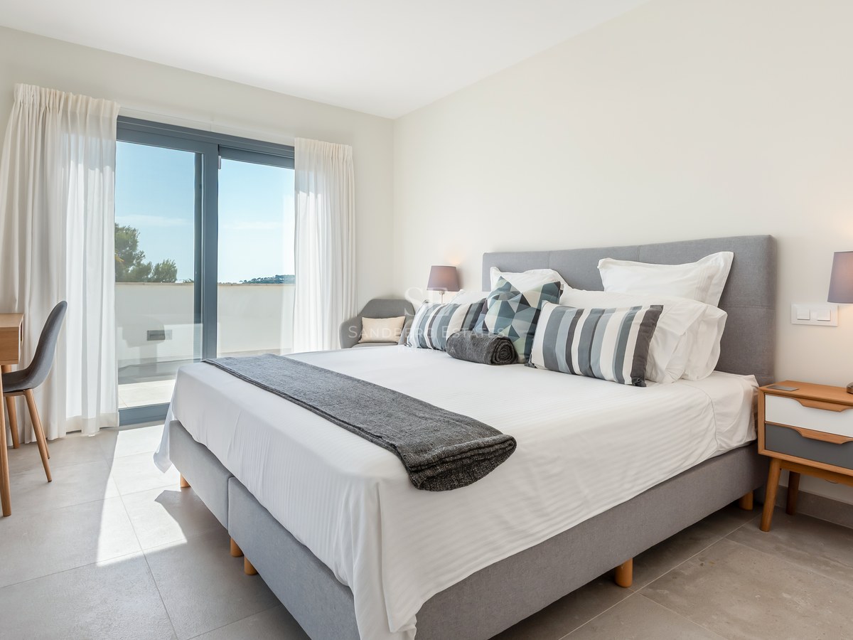 Bright bedroom with gray upholstered bed, wooden desk, and sliding glass doors to a sunny terrace.