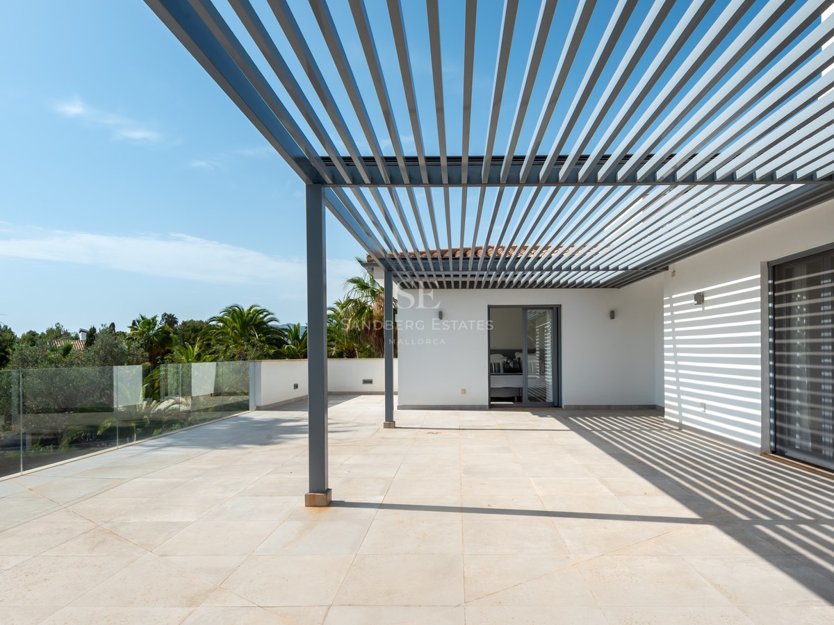 Large sun-drenched terrace with a gray metal pergola, glass railings, and views of palm trees under a blue sky.