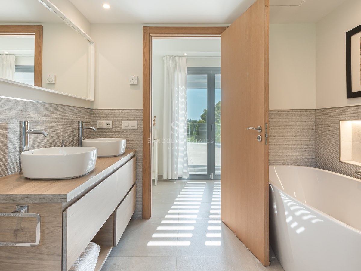 Modern bathroom featuring double vessel sinks on a wooden vanity, a large bathtub, and textured beige wall tiles.