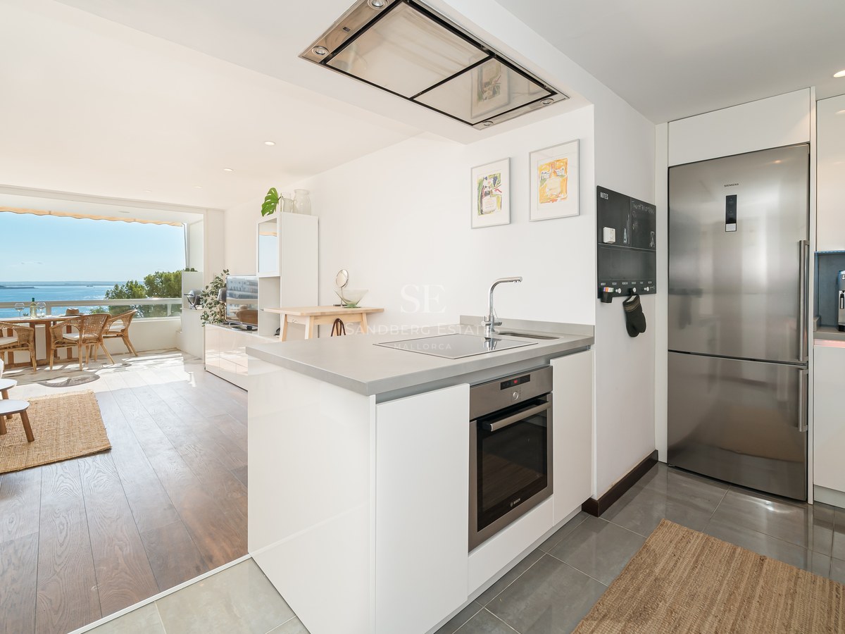 Cocina blanca moderna con electrodomésticos de acero inoxidable, salón y terraza con vistas al mar Mediterráneo.