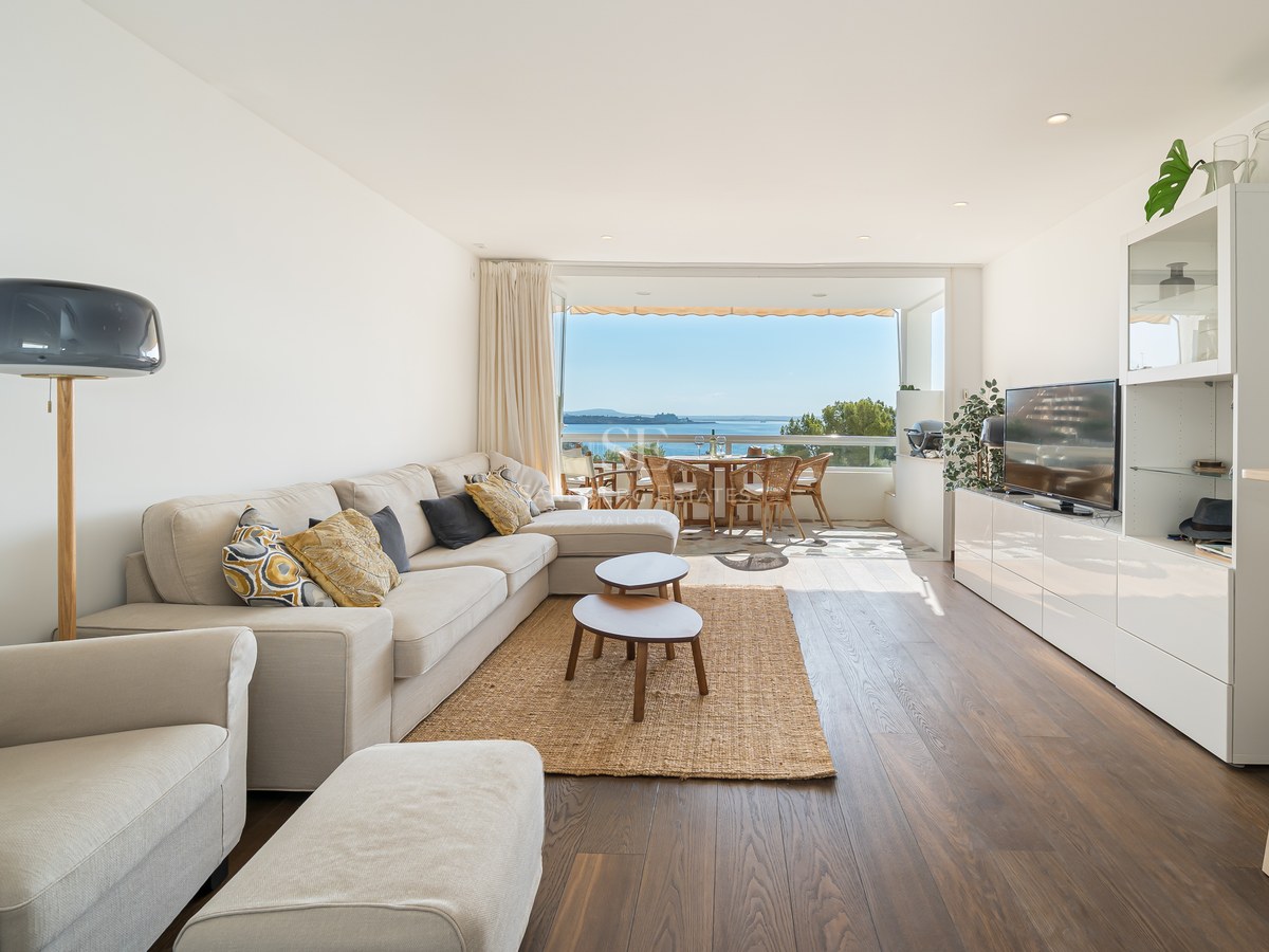 Salón luminoso con sofá grande, suelos de madera y puertas de cristal hacia terraza con vistas al mar.