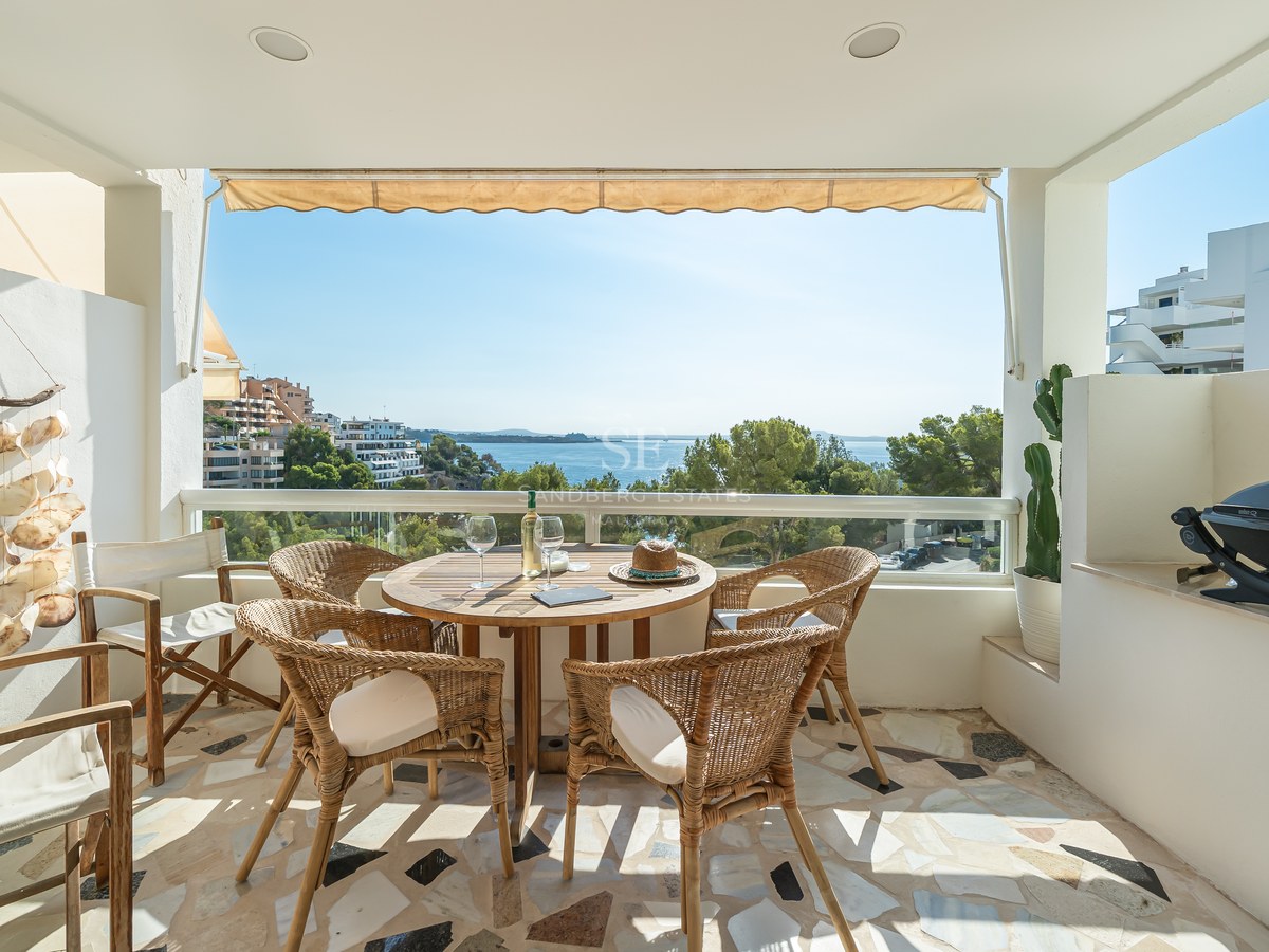 Terraza cubierta con mesa de comedor de madera, sillas de mimbre y barandilla de cristal con vistas al mar.