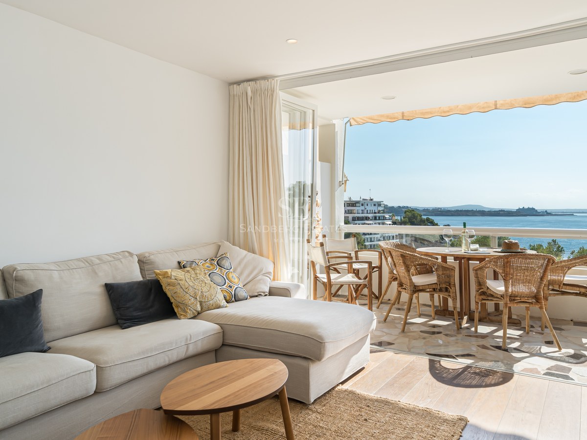 Salón interior con sofá beige que da a una terraza soleada con comedor y vistas al mar Mediterráneo.