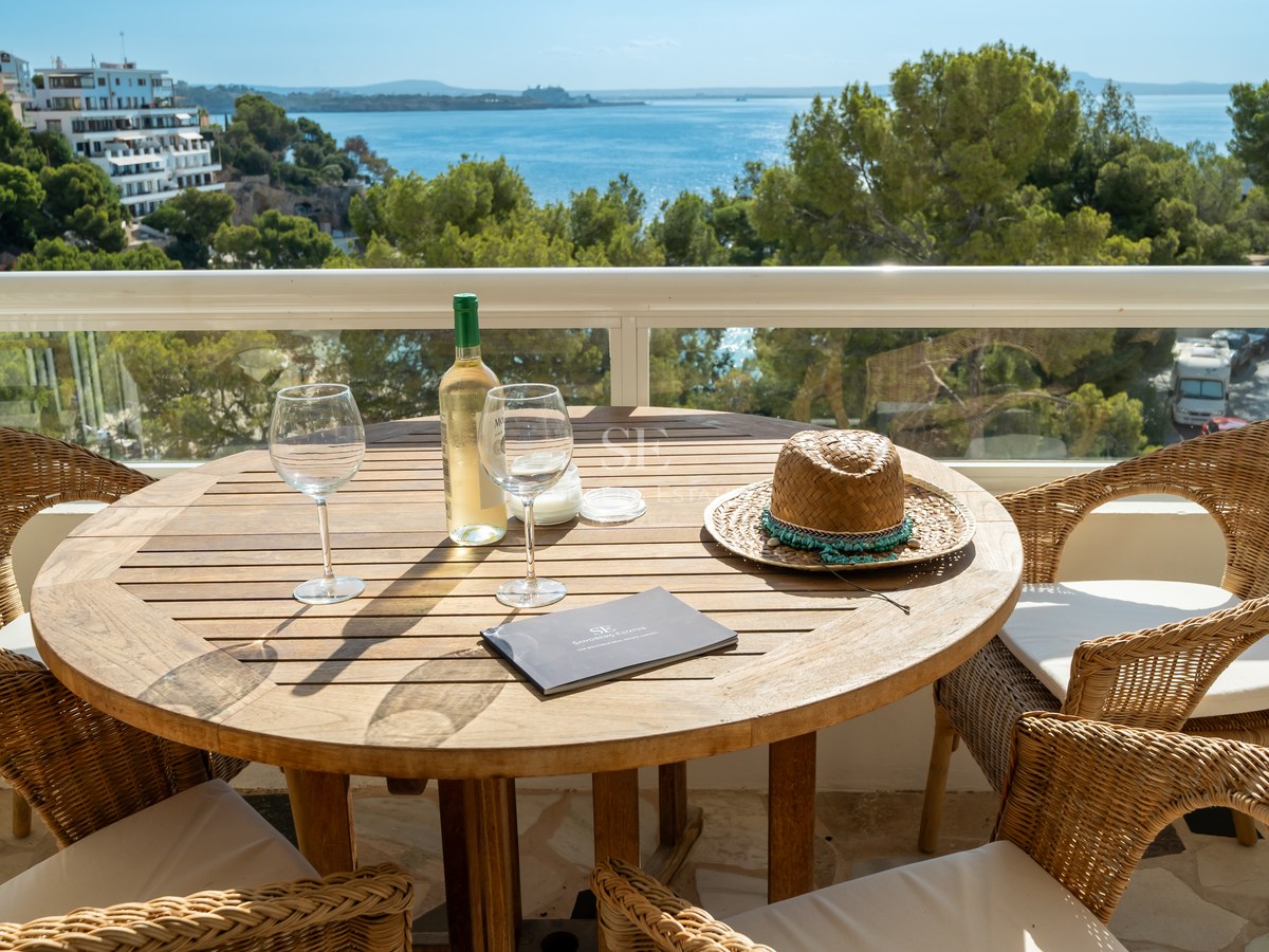 Terraza elevada con mesa de madera, sillas de mimbre y vistas al mar azul entre pinos mediterráneos.