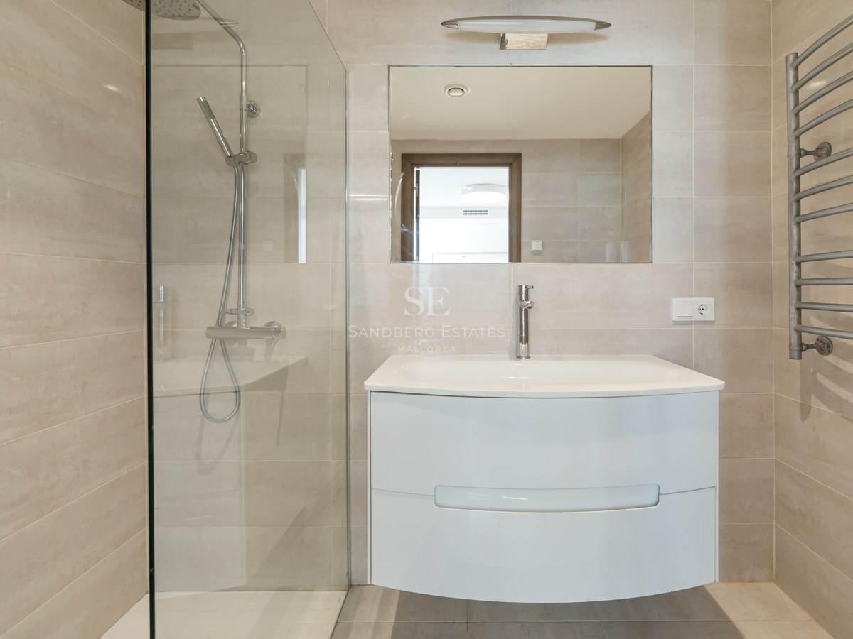 Baño moderno con azulejos beige, ducha con mampara de cristal, lavabo suspendido blanco y toallero calefactable.