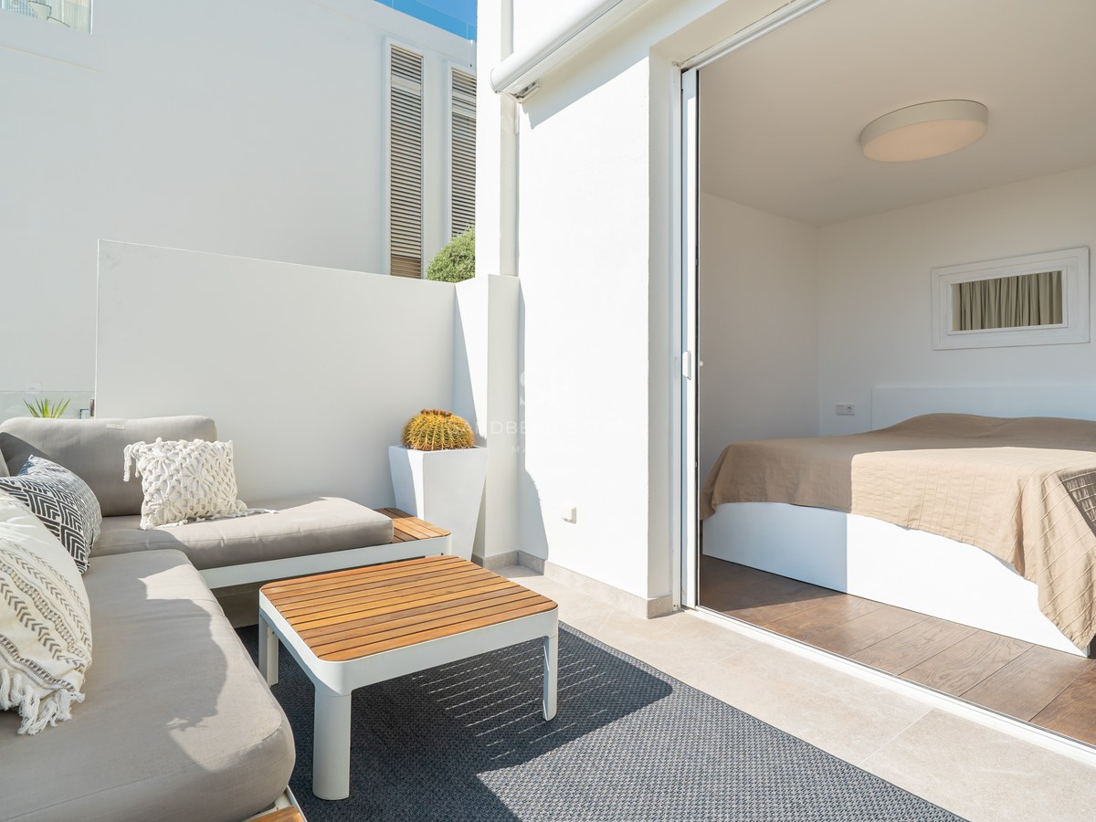 Dormitorio moderno abierto a una terraza soleada con sofás grises, mesa de madera y paredes blancas minimalistas.