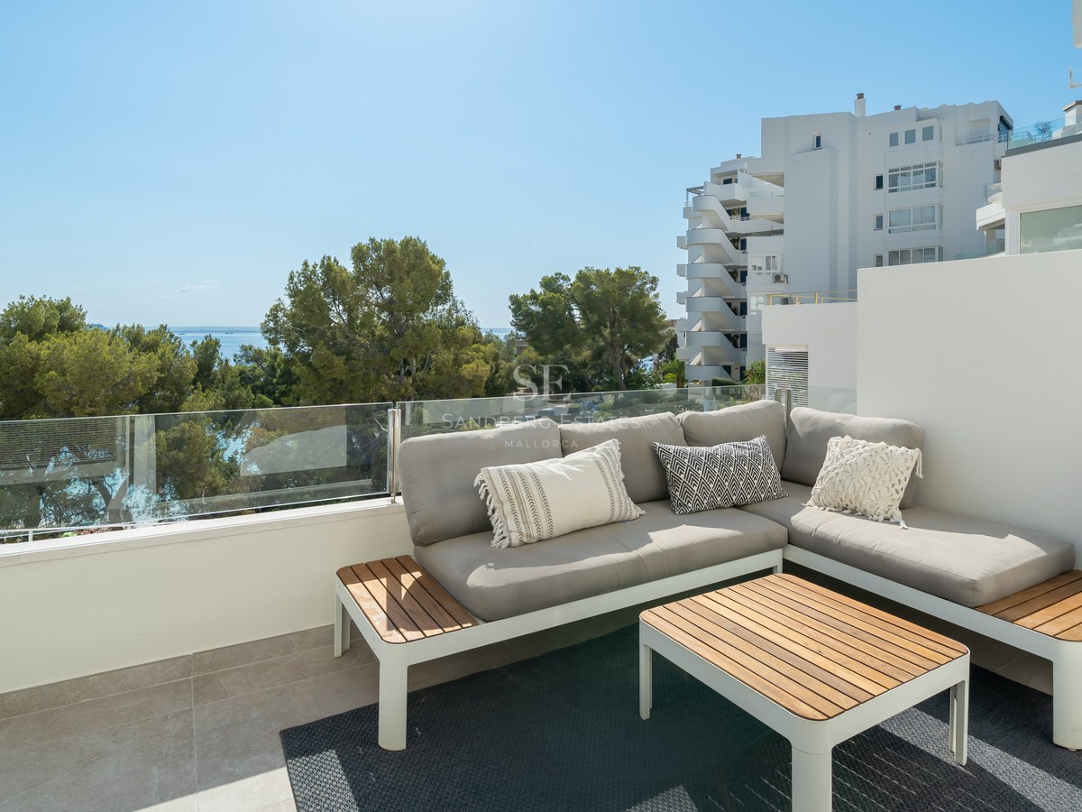 Terraza con sofá gris en L, mesa de madera y barandilla de cristal con vistas a pinos y al mar Mediterráneo.