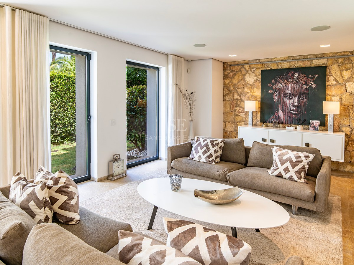 Salon moderne avec mur en pierre naturelle, canapés taupe et portes vitrées s'ouvrant sur un jardin verdoyant.