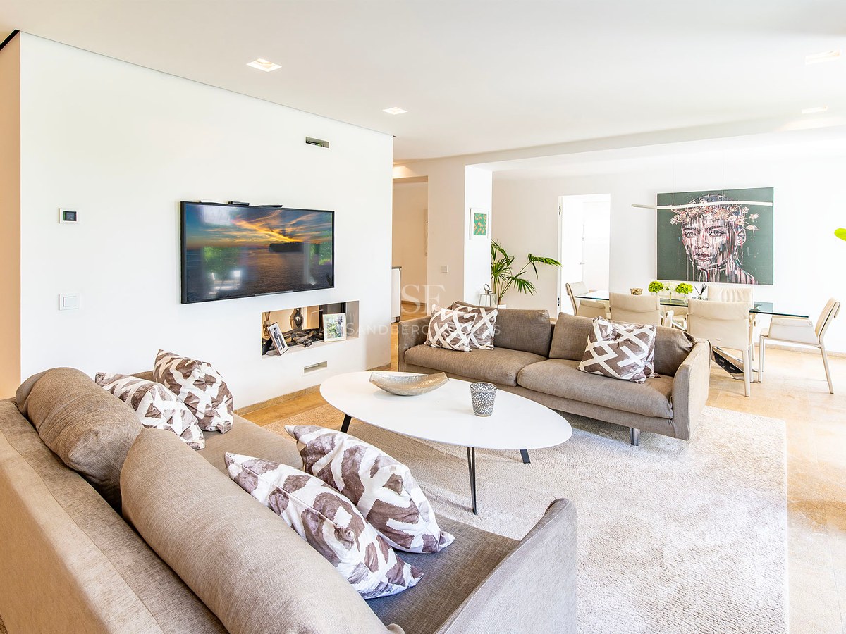 Vaste salon moderne avec canapés taupe, table basse ovale blanche et salle à manger avec œuvre d'art en arrière-plan.