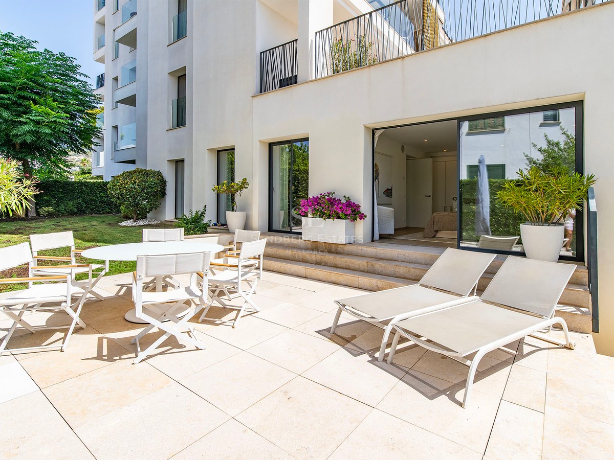 Terrasse ensoleillée avec coin repas blanc et chaises longues à côté d'un jardin et d'un immeuble moderne.
