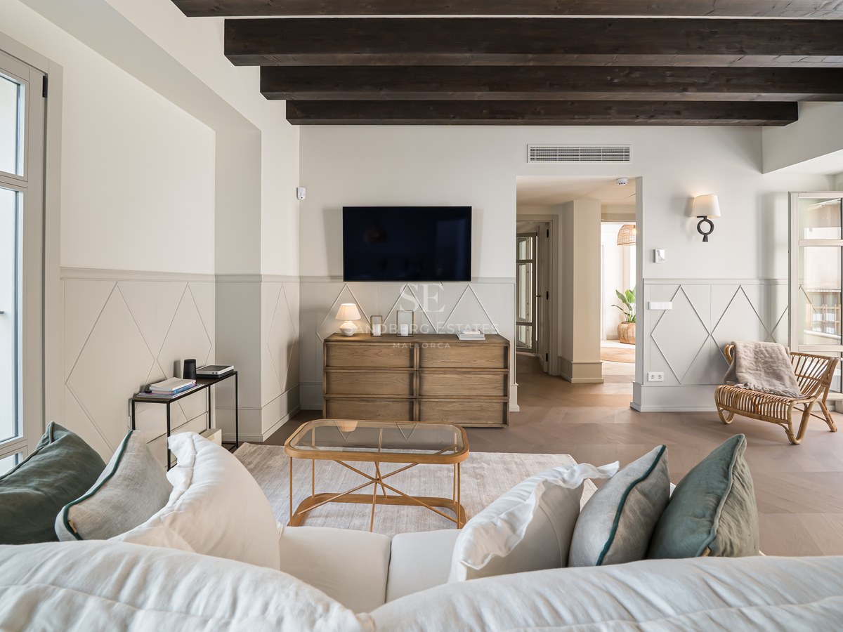 Bright living room featuring dark exposed ceiling beams, decorative wall paneling, and large windows leading to a balcony.