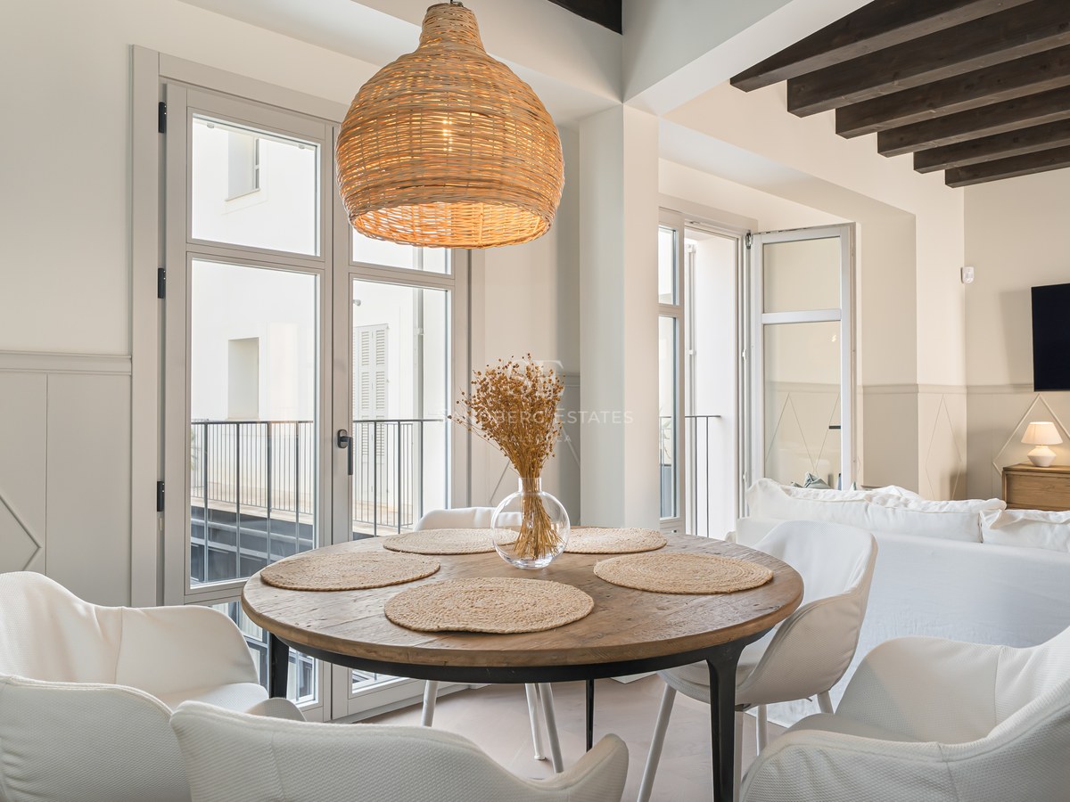 Modern round wooden dining table with white chairs, wicker pendant light, and dark exposed ceiling beams.