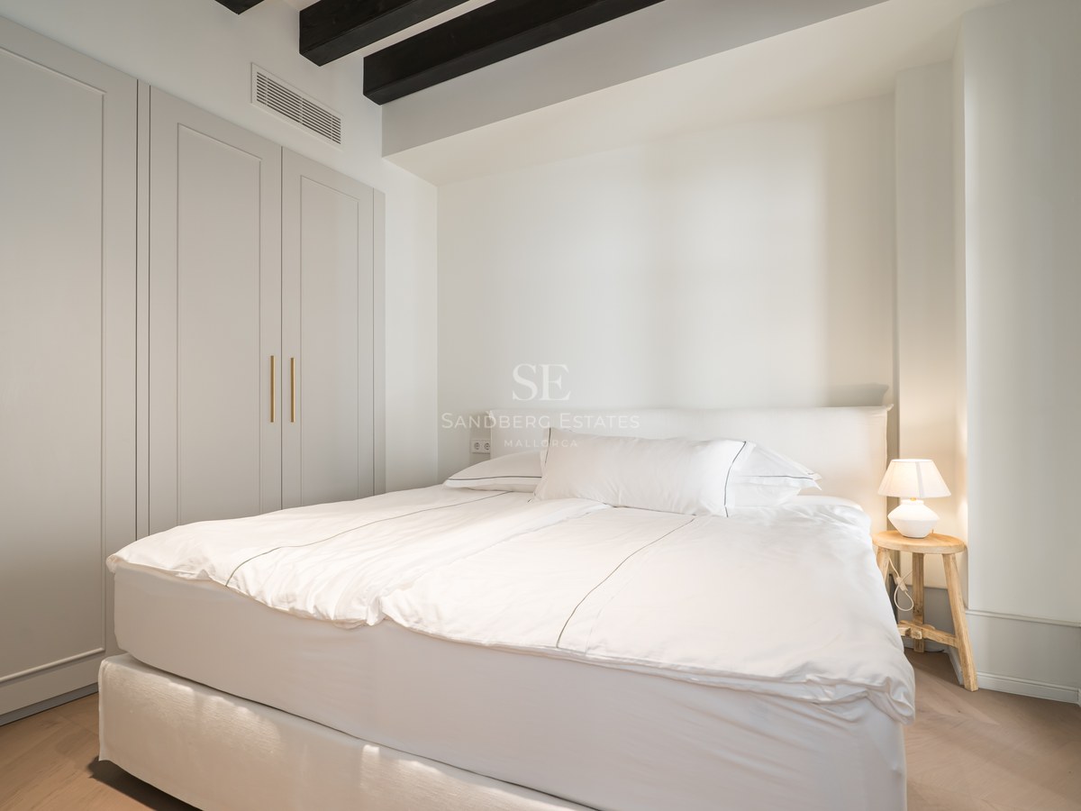 Bright bedroom with white linens, dark wood ceiling beams, built-in wardrobes, and light oak flooring.