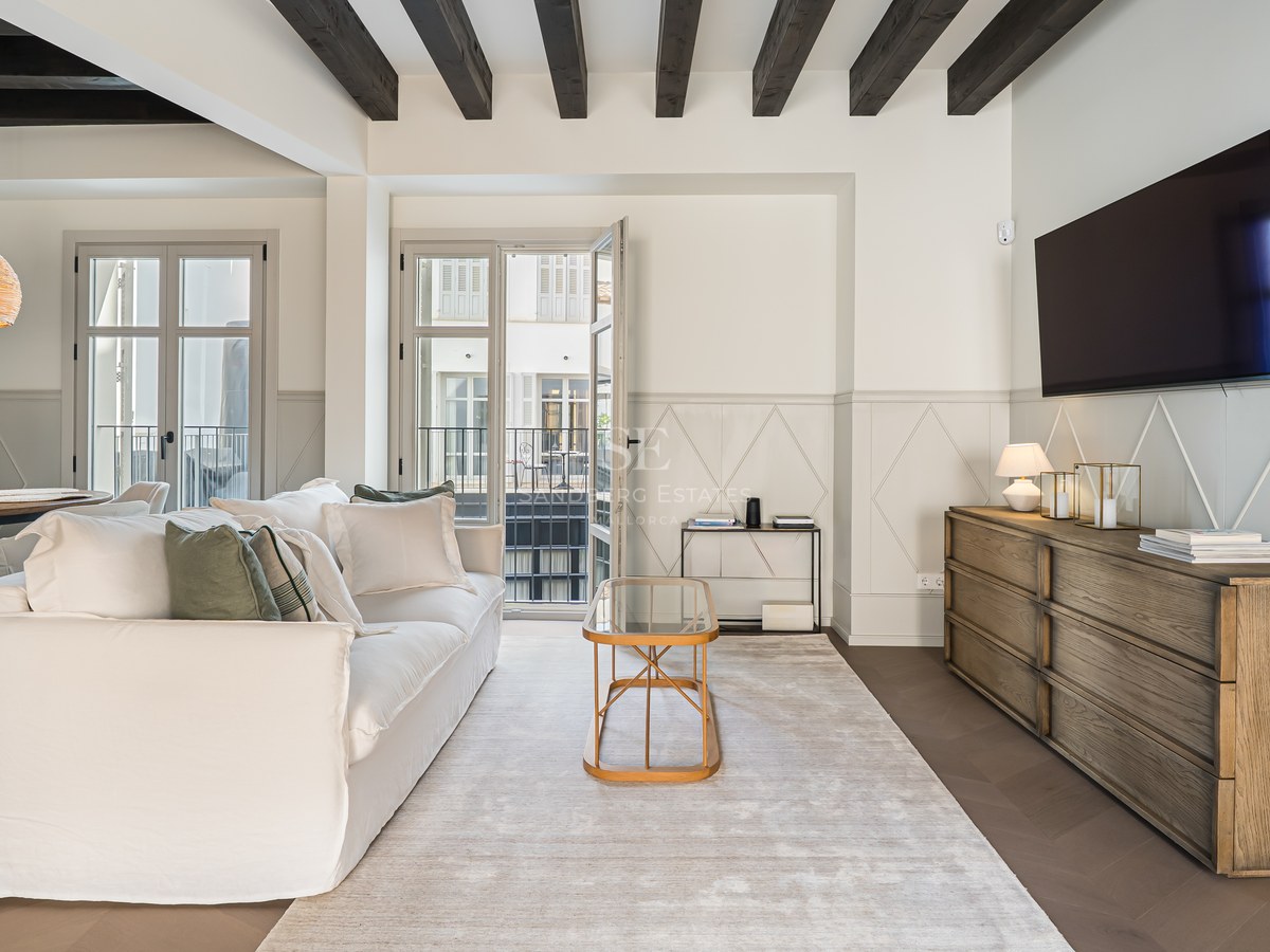 Bright living room featuring a white sofa, dark wooden ceiling beams, a wooden sideboard, and French balcony doors.