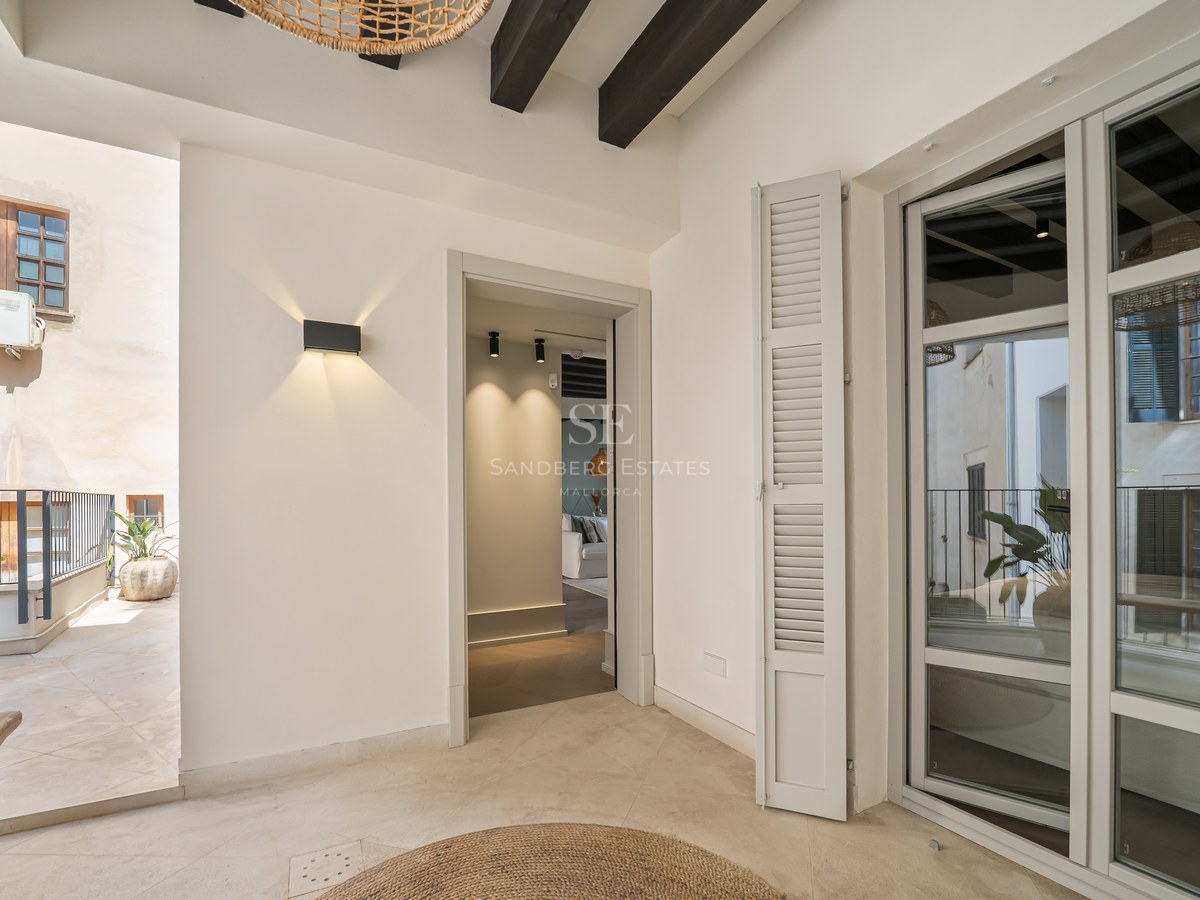 Covered terrace area with cream stone floors, dark ceiling beams, and a view into a modern interior.