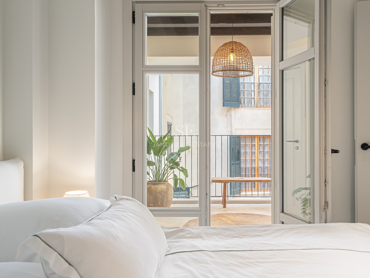 A bright bedroom with white linens looking through open glass doors to a balcony with a woven lamp and plants.