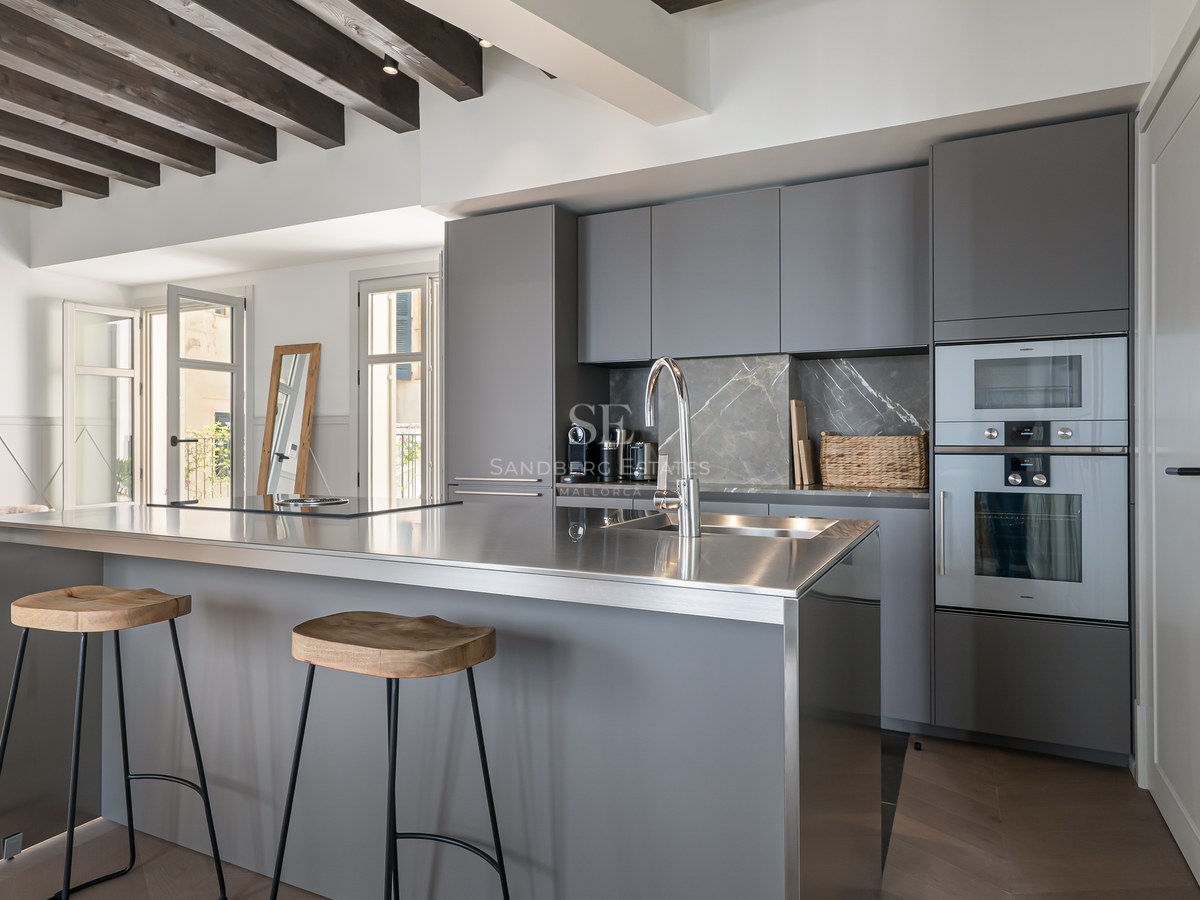 Modern kitchen featuring grey cabinetry, a stainless steel island with bar stools, and exposed wooden ceiling beams.