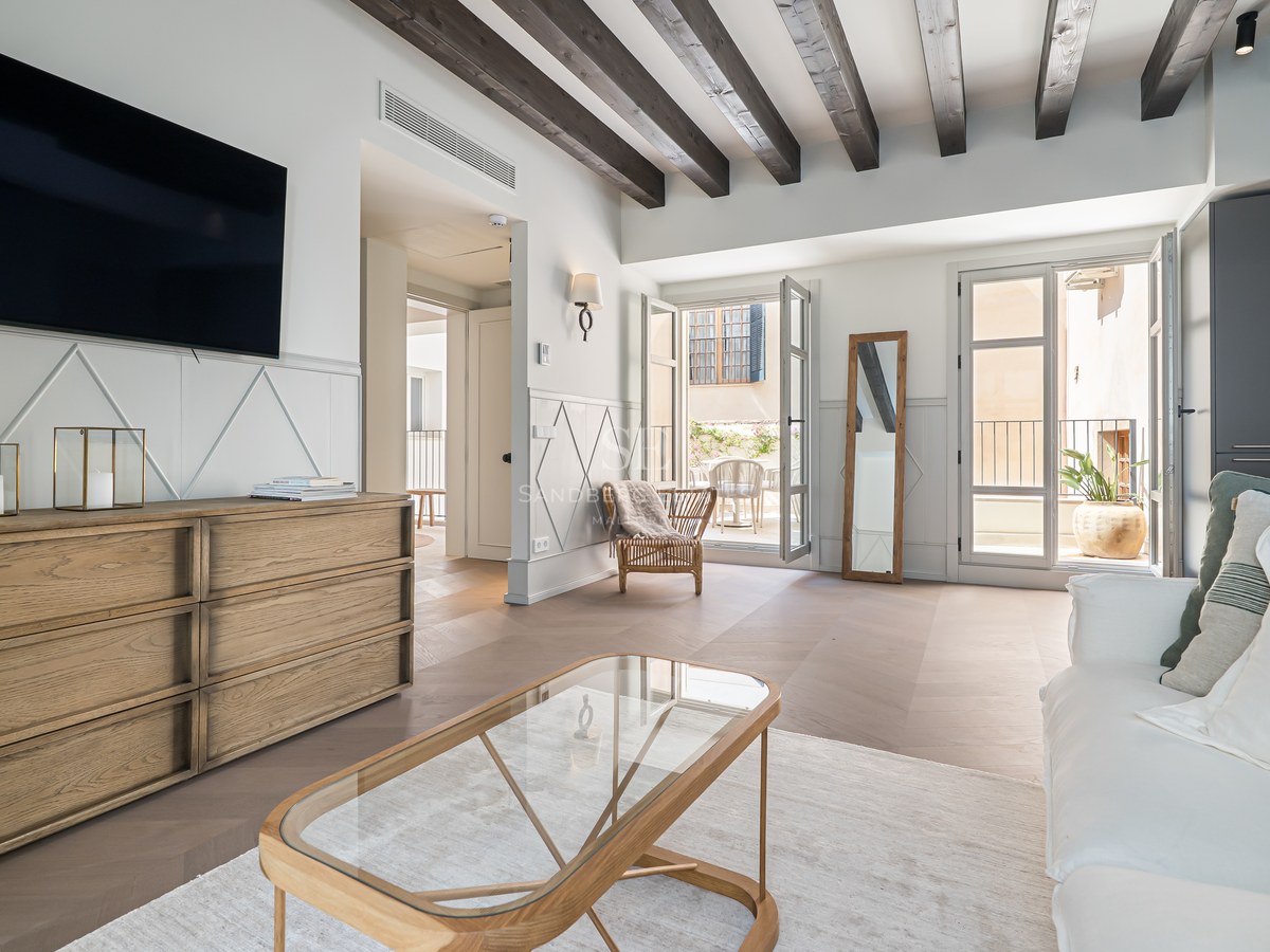 Bright contemporary living room with dark wooden ceiling beams, oak furniture, and doors opening to a balcony.