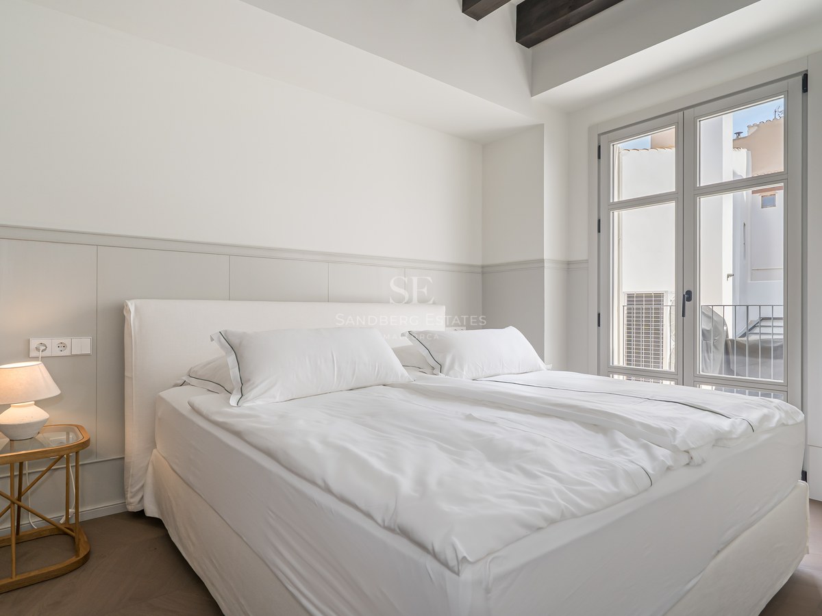 Bright bedroom featuring white linens, dark wooden ceiling beams, and a French window opening to a Juliet balcony.