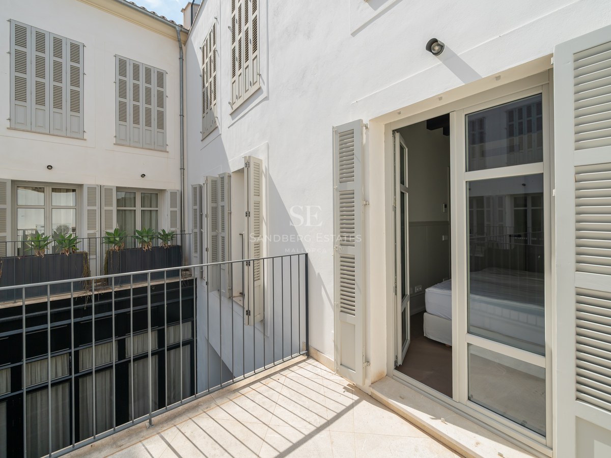 Private terrace with light stone flooring, black iron railings, and grey wooden shutters on a clean white facade.