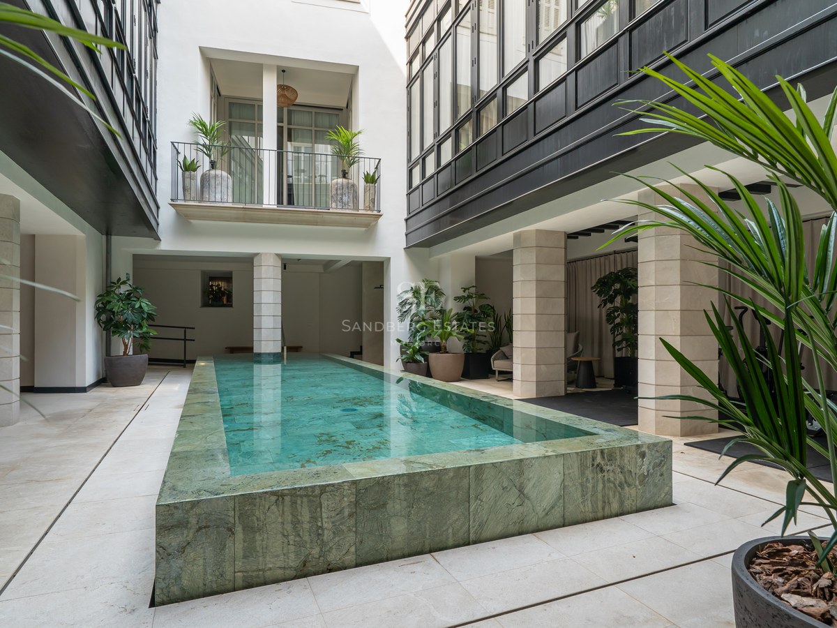 A rectangular raised lap pool made of green natural stone in a white-walled courtyard with columns and palms.
