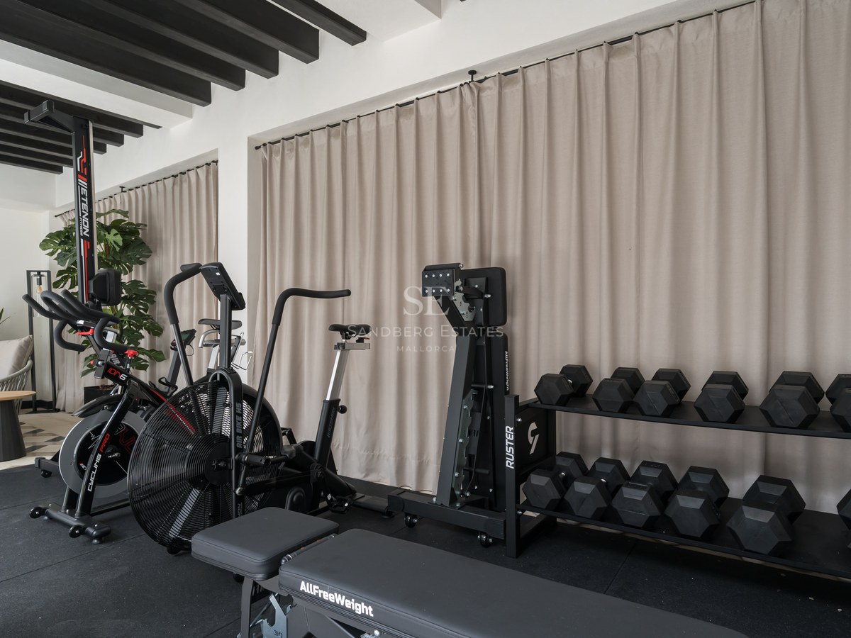 Modern home gym with stationary bikes, weight rack, and workout bench against beige curtains and black ceiling beams.