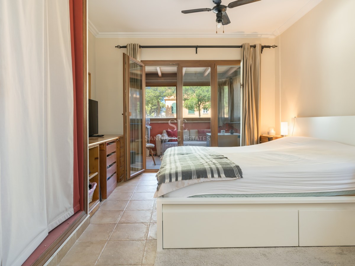 Dormitorio con suelos de terracota, cama blanca y puertas acristaladas que dan a una terraza amueblada.