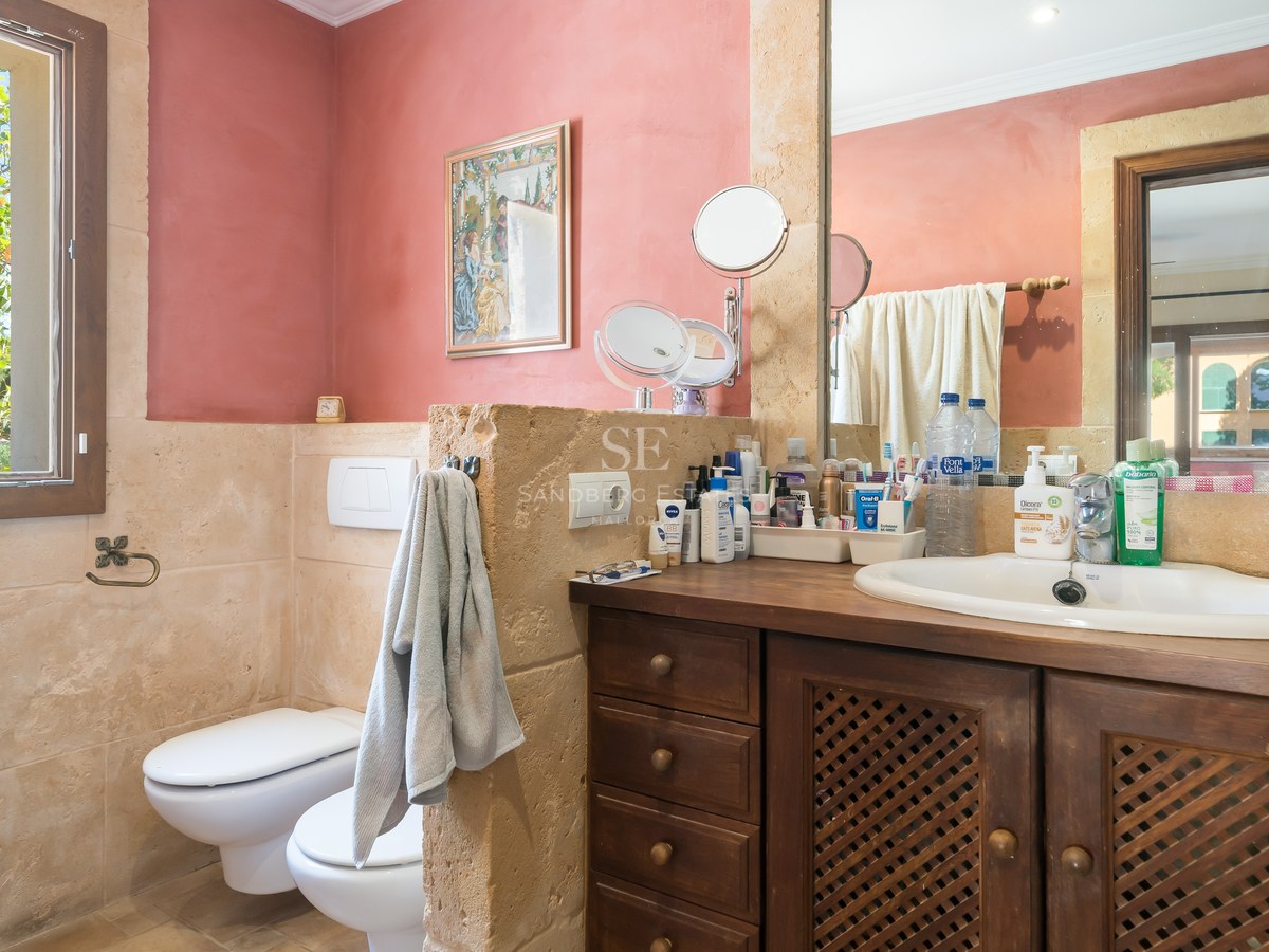 Cuarto de baño con paredes de piedra natural, mueble de madera oscura, inodoro y bidé, con ventana al jardín.