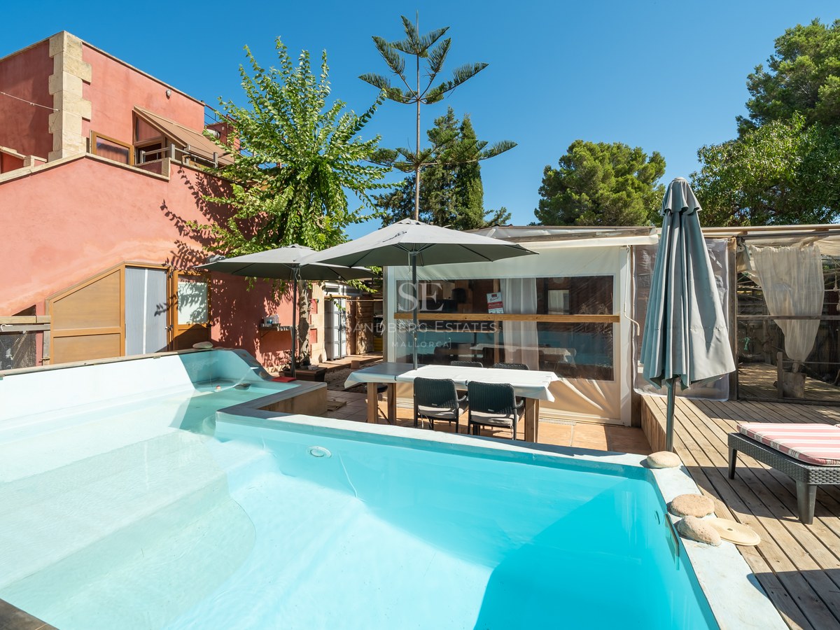 Piscina turquesa junto a terraza de madera con tumbonas y fachada de terracota bajo un cielo despejado.