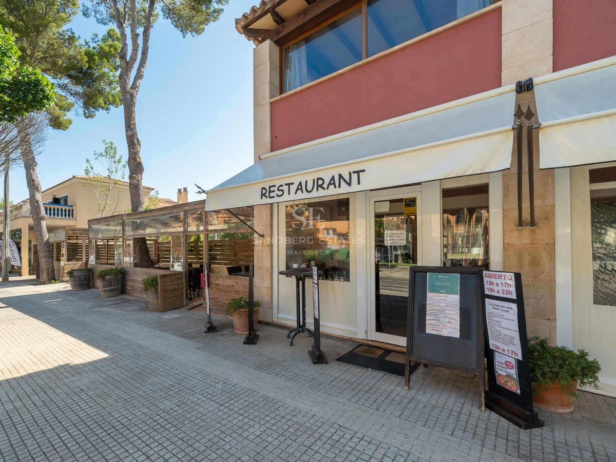 Vista exterior de un restaurante con paredes de piedra, toldos blancos y una acera arbolada bajo un cielo despejado.