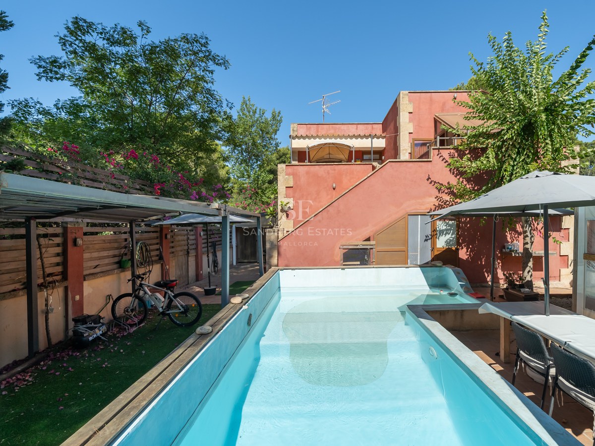 Piscina rectangular turquesa en un jardín privado con una casa color terracota y zona de comedor bajo sombrilla.