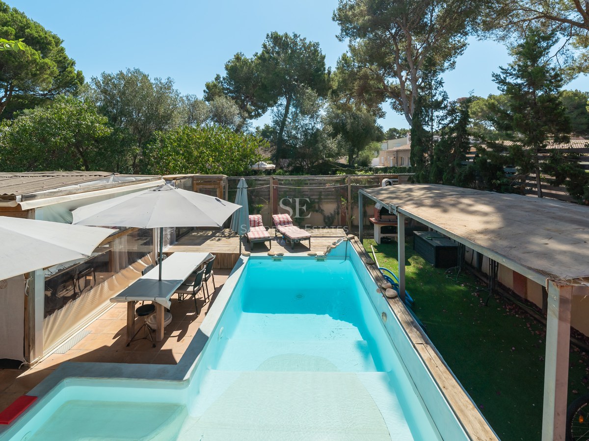 Piscina rectangular turquesa rodeada de tarima de madera, comedor exterior y árboles bajo un cielo azul despejado.