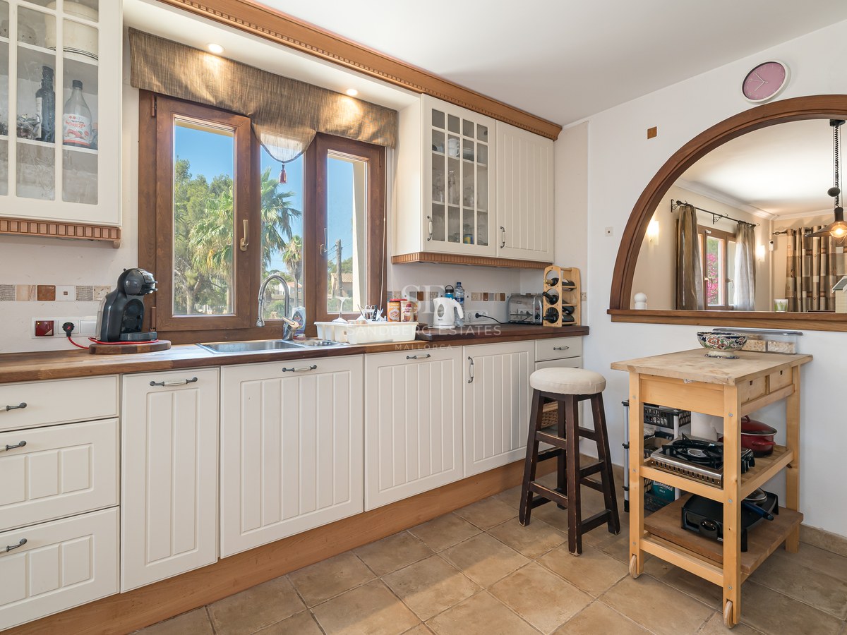Cocina crema con encimeras de madera, suelo de terracota y ventana de arco hacia el comedor y jardín.