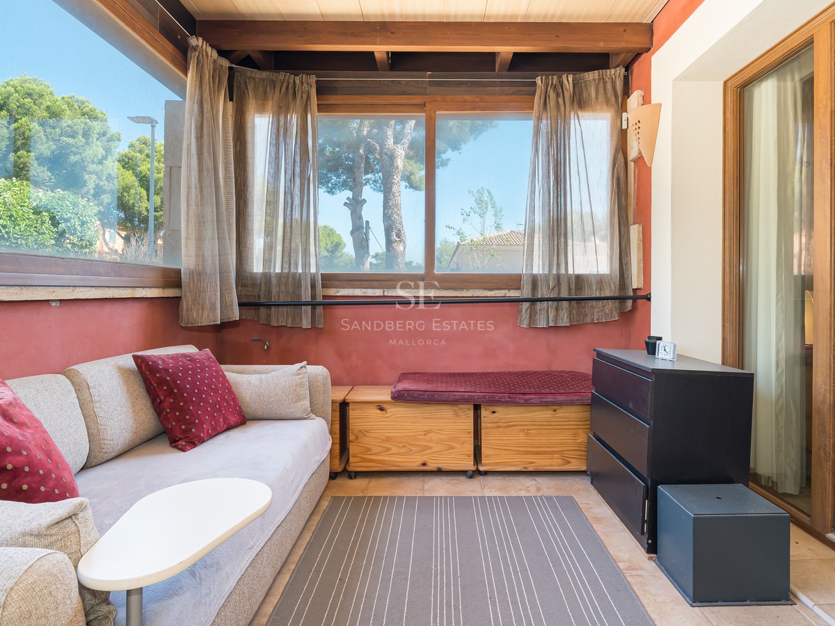 Sala de estar iluminada con sofá beige, vigas de madera, paredes de terracota y grandes ventanas con vistas al jardín.