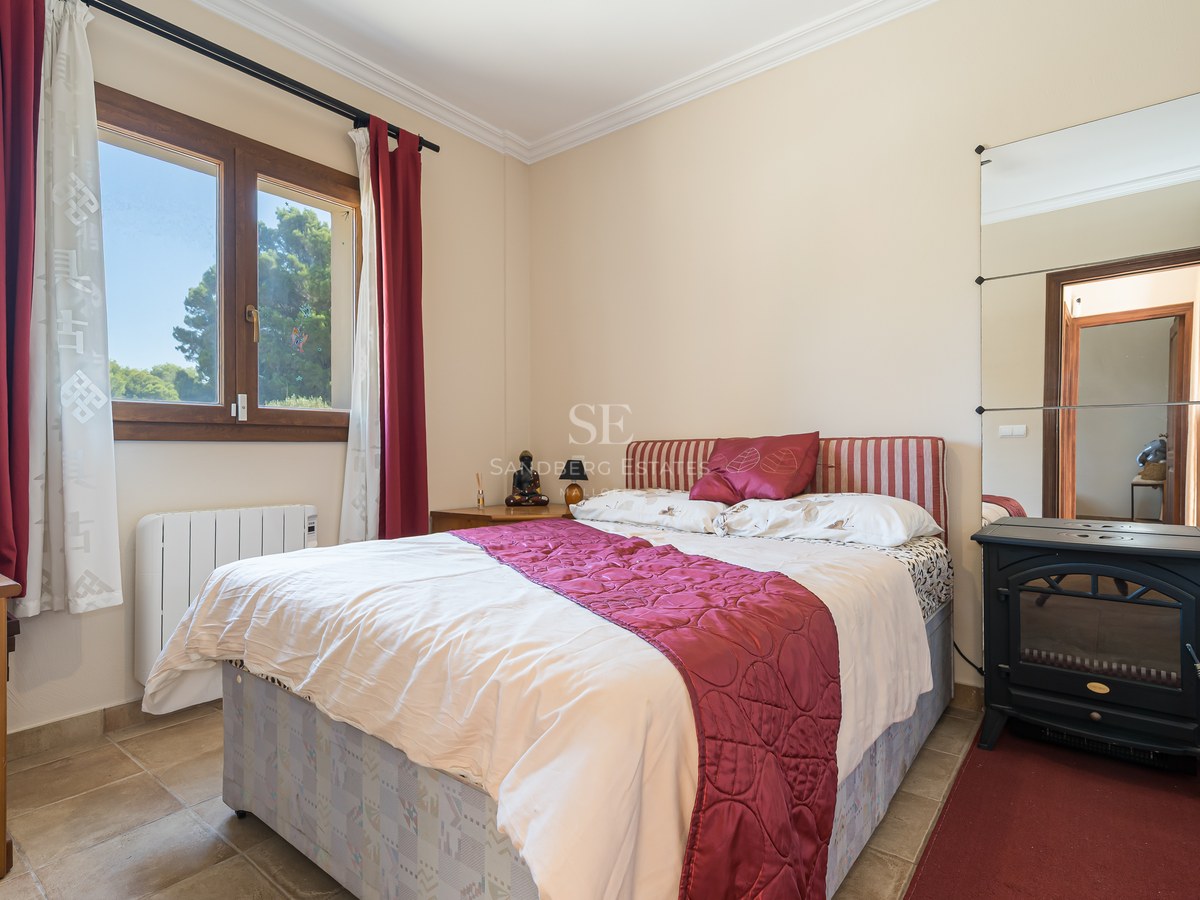 Dormitorio con cama doble, textiles granates, ventana de madera con vistas a pinos y estufa decorativa negra.