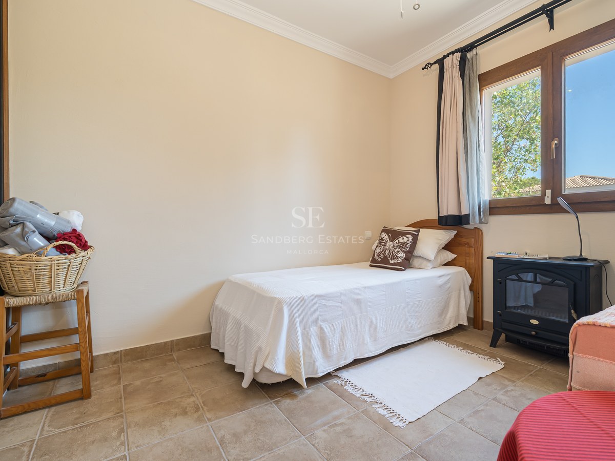 Pequeño dormitorio con cama individual, suelo de baldosas, ventana de madera y estufa decorativa.