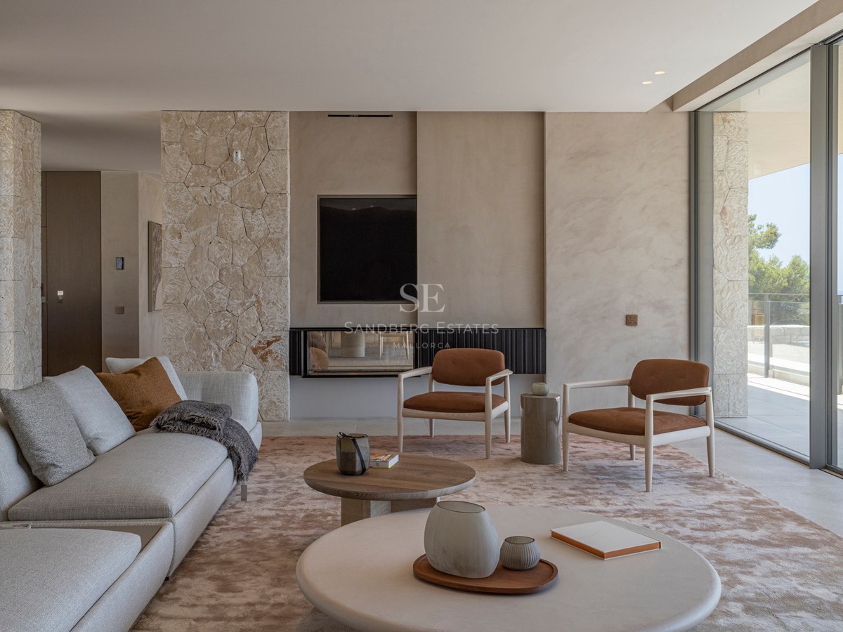 Salon moderne avec murs en pierre, canapé beige, fauteuils terre cuite et grandes baies vitrées vue mer.