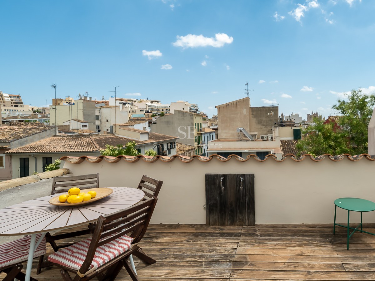Terraza en la azotea con suelo de madera y zona de comedor con vistas a los tejados de una ciudad mediterránea.