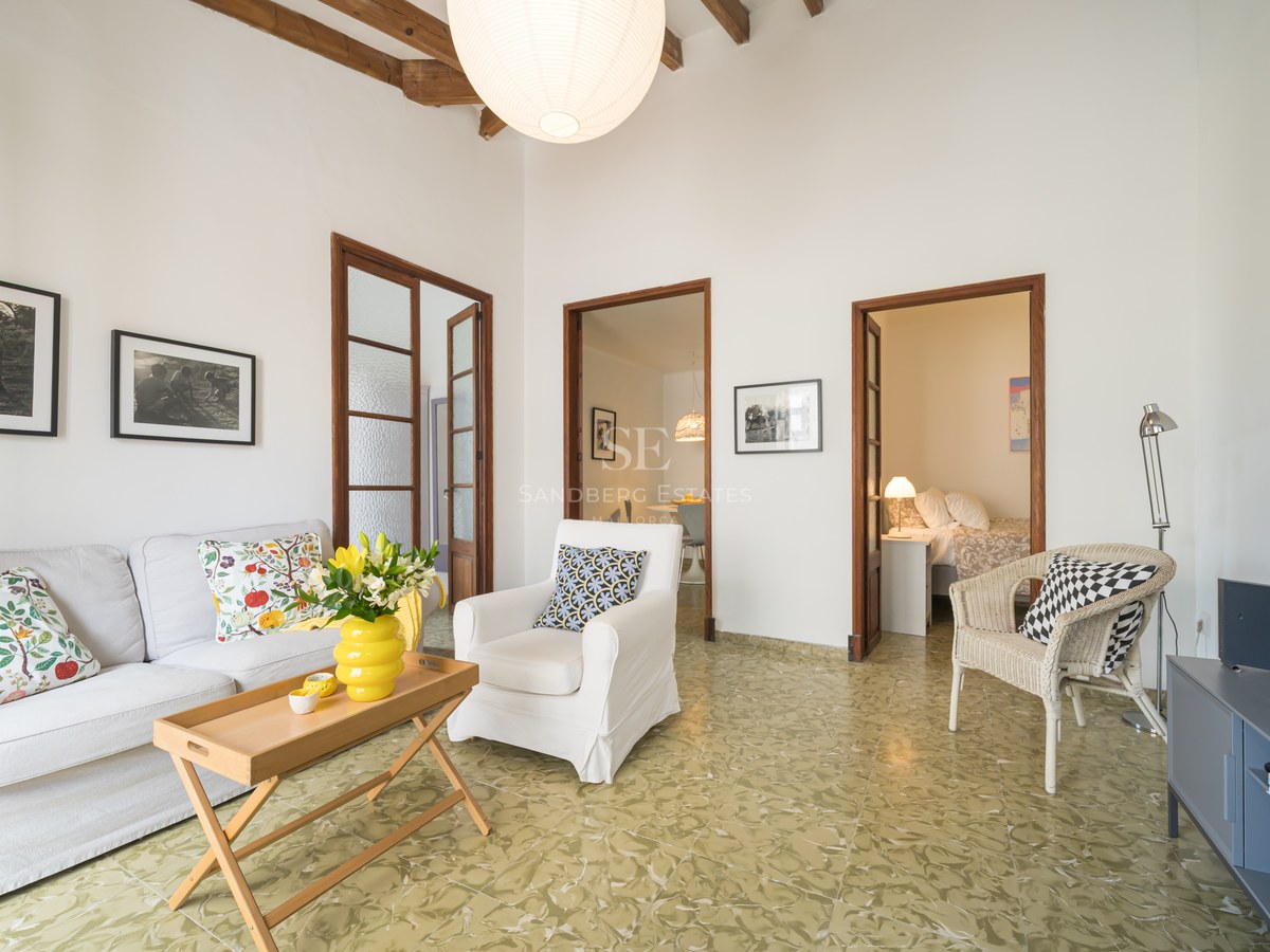 Salón luminoso con sofá blanco, vigas de madera rústicas, suelo de baldosas verdes estampadas y puertas a otras estancias.